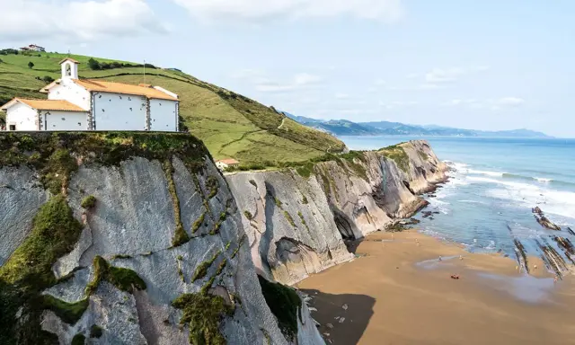 Ermita blanca sobre acantilados en la playa de Itzurun, con el mar Cantábrico y olas rompiendo.