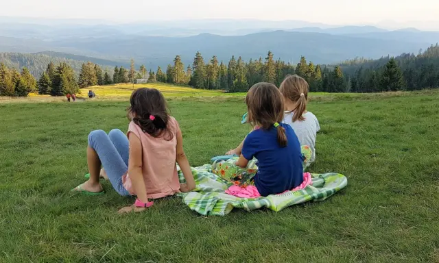 Trzy dziewczynki odpoczywają na kocu na trawie, podziwiając widok gór. W tle widać ludzi idących szlakiem.