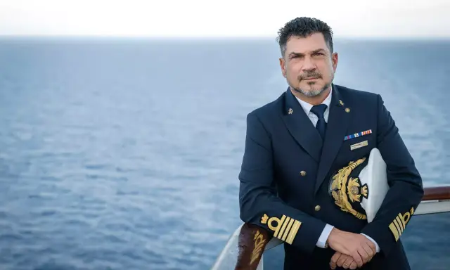 Il comandante MSC Splendida, con la sua uniforme blu scuro e il cappello dorato, posa con il mare calmo sullo sfondo.
