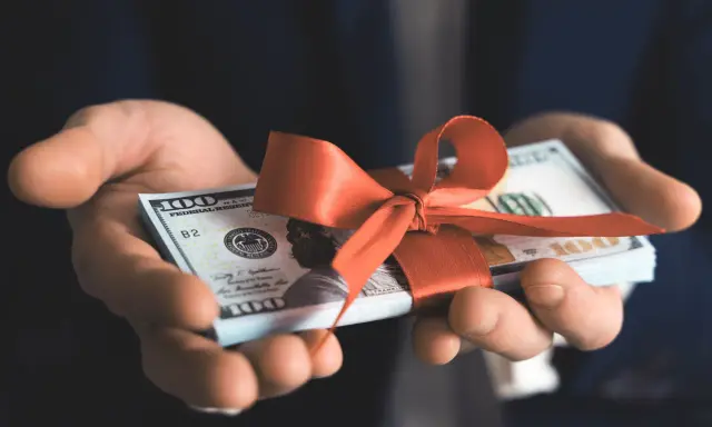 Dłonie trzymają plik banknotów 100-dolarowych przewiązany czerwoną wstążką. To pieniądze zamiast prezentów na urodziny, praktyczny i cenny podarunek.