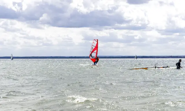 Czerwony żagiel do windsurfingu na wodzie, surfer w akcji. W tle inni windsurferzy i chmury.