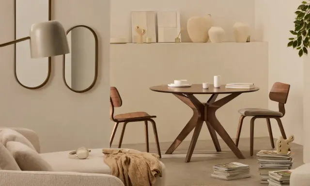 Dos espejos ovalados cuelgan sobre un sofá beige. A la derecha, una mesa redonda de madera con sillas y un jarrón.