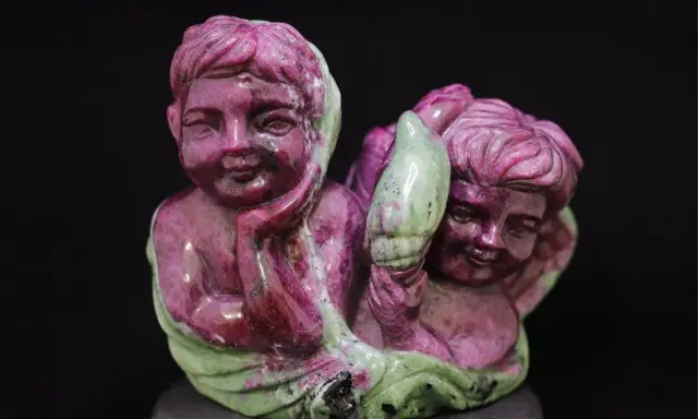 Due putti scolpiti in pietra, uno con la mano sul mento, sembrano abbracciare un elemento verde, forse un fiore. La pietra, un rubino verde, dona un tocco di magia.