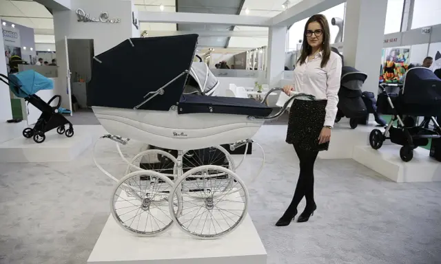 Elegancka kobieta prezentuje najdroższe wózki dziecięce, w tym luksusowy model Silver Cross z białym koszem i czarną gondolą.
