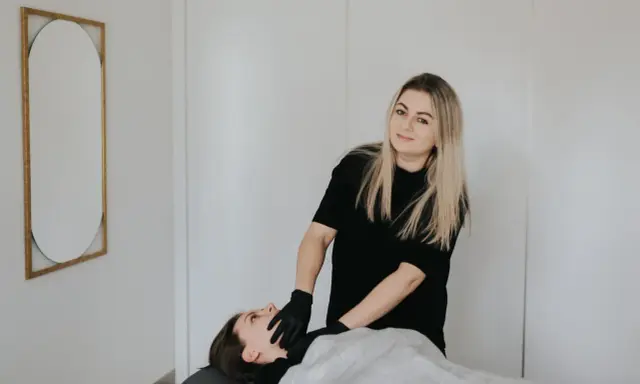 Terapeuta w czarnych rękawiczkach wykonuje zabieg rehabilitacji głosu u leżącej pacjentki.