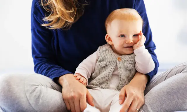 Uśmiechnięte dziecko w sweterku, które zaczyna siadać, siedzi na kolanach mamy.