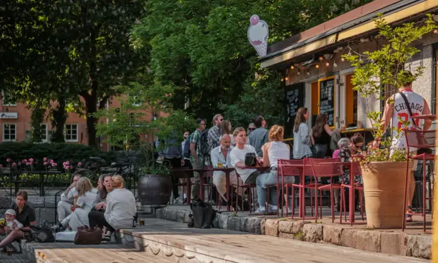 Folk njuter av solen på uteserveringar i Stockholm. En glasskylt syns ovanför en servering.