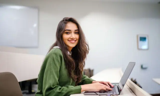 Uśmiechnięta studentka pracuje na laptopie w sali wykładowej. Jej status ucznia jest widoczny w otoczeniu i skupieniu na nauce.