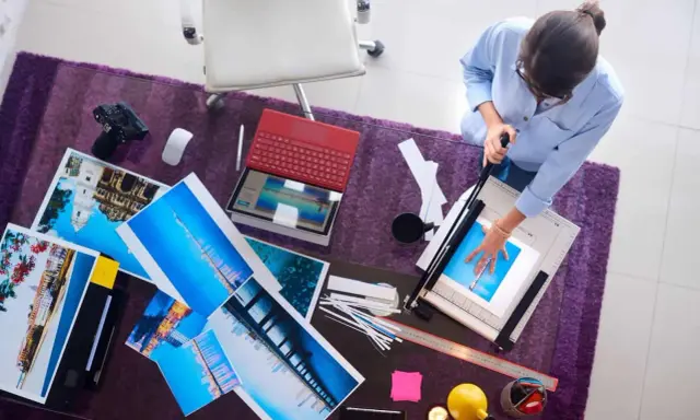 Kobieta przycina wydruki wykonane techniką druk lateksowy. Na stole leżą zdjęcia, laptop i aparat.