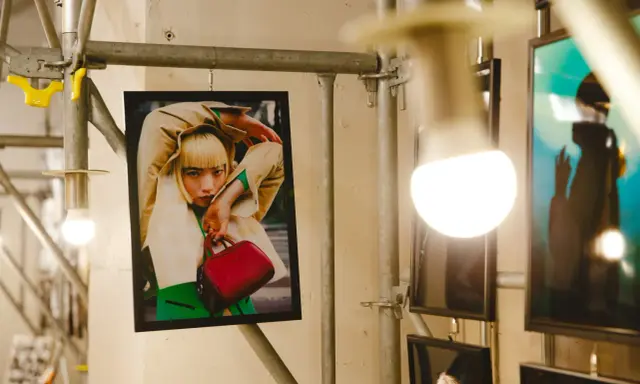 Plakat z kobietą w beżowym płaszczu i zielonym swetrze, trzymającą czerwoną torebkę. Stylizacja przypomina modę z **Pezet Tokyo**.
