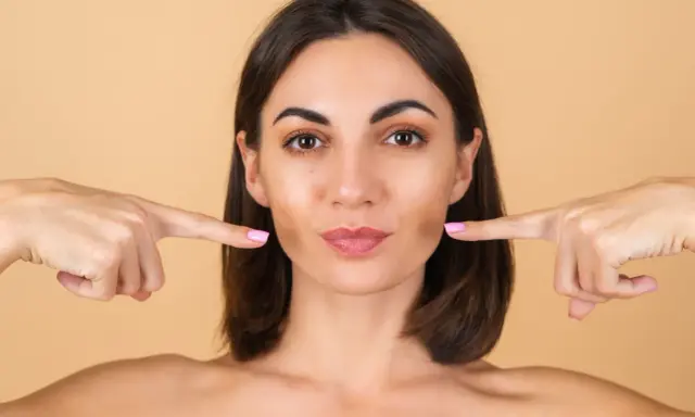 Doigts pointés vers les joues, une femme montre comment repulper son visage pour un look plus jeune et plein.