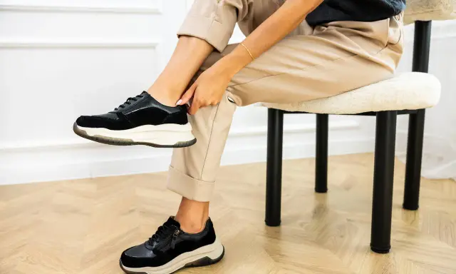 Kobieta w beżowych spodniach i czarnych sneakersach na grubych podeszwach. Pokazuje, jak szybko wysuszyć buty, by znów cieszyć się komfortem.