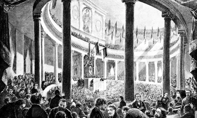 Versammlung der Frankfurter Nationalversammlung in der Paulskirche. Viele Männer in Anzügen und Hemden sitzen und stehen in einem großen, säulenreichen Gebäude.
