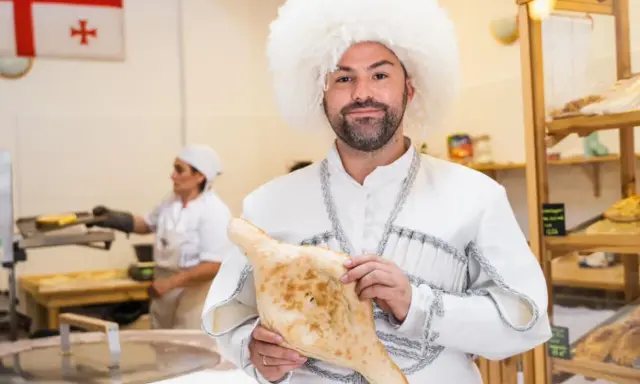 Mężczyzna w tradycyjnym stroju i czapce z owczej wełny trzyma chleb gruziński. W tle widać flagę Gruzji i piekarnię.