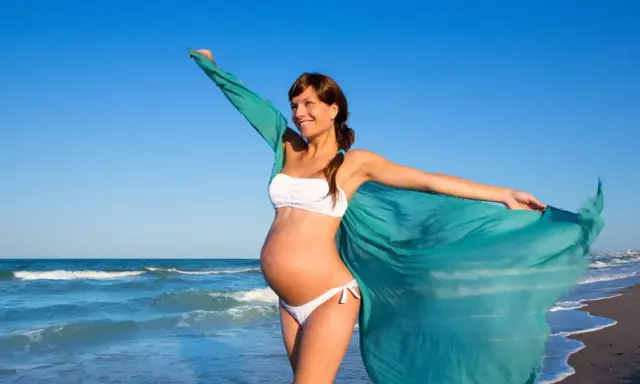 Donna incinta in bikini bianco sulla spiaggia, con un velo turchese che svolazza al vento. Il mare sullo sfondo.