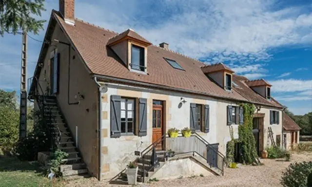 Une maison de campagne simple avec des volets gris, une cheminée et des escaliers extérieurs sous un ciel bleu.