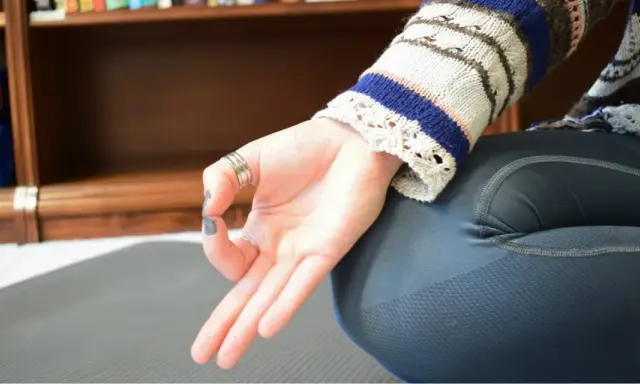 En hand utför en mudra, en gest som används i yoga och meditation. Bakom syns en bokhylla med yogamattor.