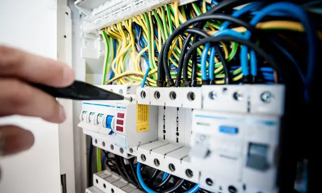 Ręka z narzędziem przy bezpiecznikach. Widać kolorowe przewody i moduły, tworzące prostą instalację elektryczną.
