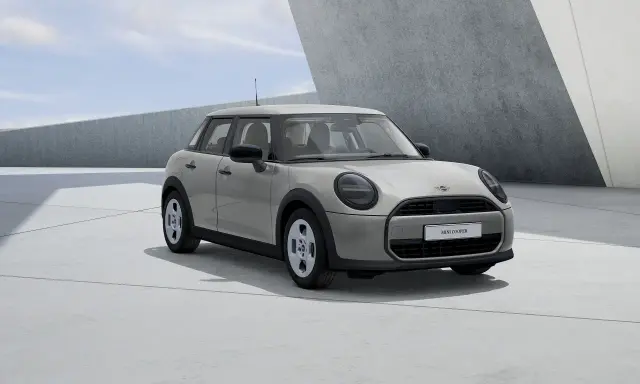 Mini Cooper 5 puertas en color gris claro, con llantas plateadas, estacionado en una zona de concreto con arquitectura moderna.
