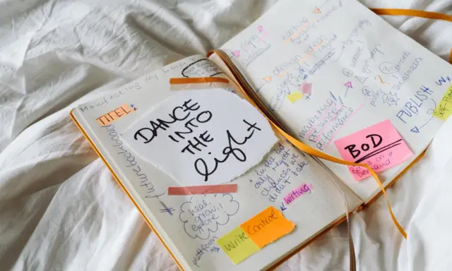 Carnet ouvert avec des notes manuscrites sur la création d'un livre, incluant "Dance into the Light" et des idées de marketing. Un nom d'auteur connu pourrait être inspiré par ces notes.