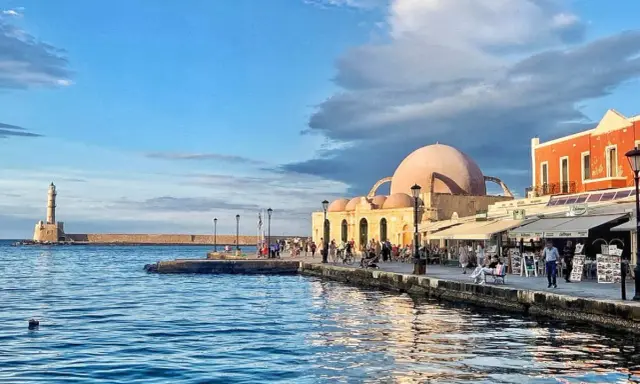 Il porto di Chania con il suo faro iconico e la moschea ottomana, bagnato da un mare blu intenso sotto un cielo nuvoloso.