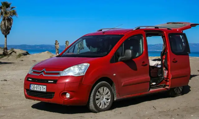 Czerwony Citroen Berlingo 1.6 HDI na plaży, z otwartymi drzwiami i bagażnikiem, na tle morza i palmy.