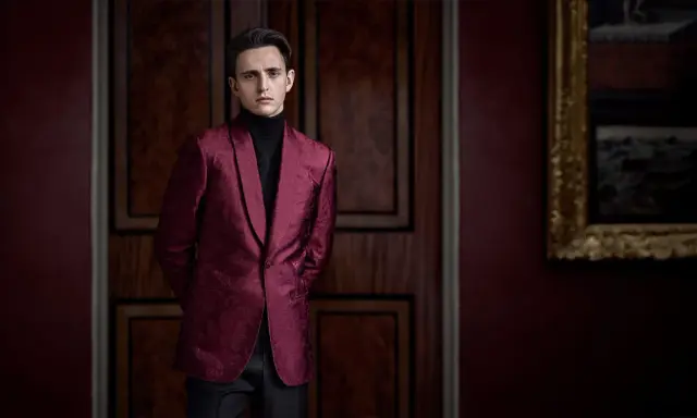 A man in a patterned burgundy jacket and black turtleneck, perfect for cocktail party attire for men, stands in a dimly lit room with ornate wooden doors.