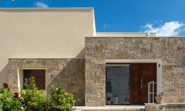 Moderne facciate in pietra grezza si alternano a intonaco chiaro, creando un contrasto materico. Un ingresso con porta in legno scuro e un giardino fiorito.