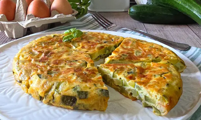 Una gustosa frittata di zucchine, dorata e soffice, servita su un piatto bianco. Ingredienti freschi come uova e zucchine sono visibili sullo sfondo.