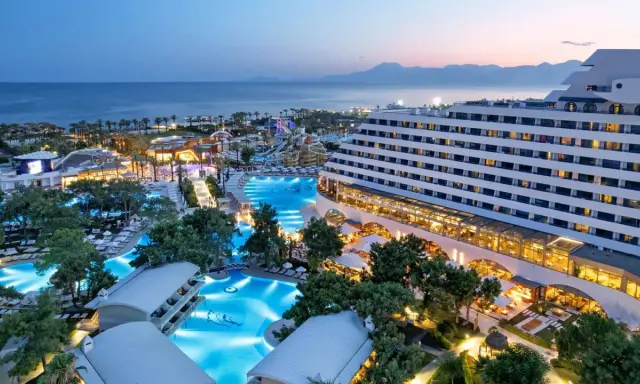 Luksusowy hotel Titanic Deluxe Lara z basenami i parkiem wodnym, oświetlony wieczorem, z widokiem na morze i góry.
