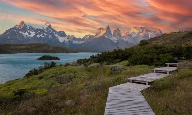 Drewniana ścieżka prowadzi przez zielone wzgórza ku turkusowemu jezioru i ośnieżonym górom. Zachód słońca maluje niebo nad **Tierra del Fuego** odcieniami różu i pomarańczy.