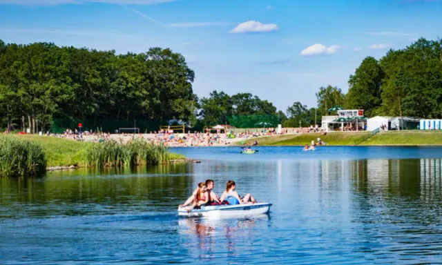 Rodzina pływa rowerkiem wodnym po jeziorze na Dolnym Śląsku, w tle plaża pełna ludzi i zielone drzewa.