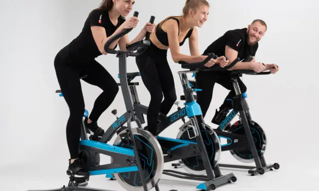 Trzy osoby ćwiczą na rowerach stacjonarnych. To świetny trening spinningowy!