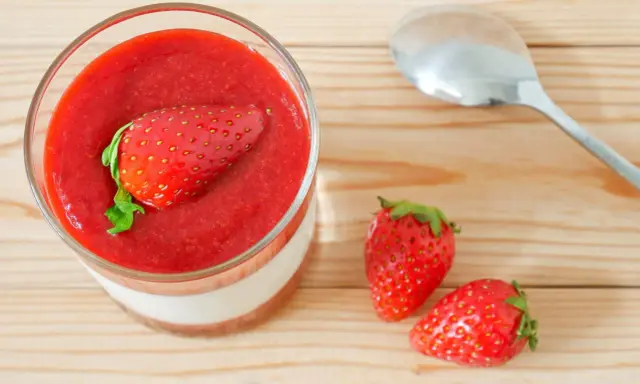 Un verre de panna cotta avec une fraise entière sur un coulis de fraise. Deux fraises fraîches et une cuillère à côté.