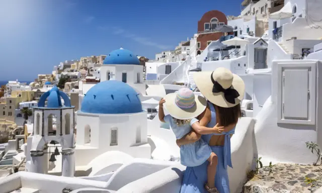 Una madre abbraccia la sua bambina, ammirando il panorama di Santorini, Grecia. Le cupole blu brillano sotto il sole, mentre le case bianche si aggrappano alla collina.