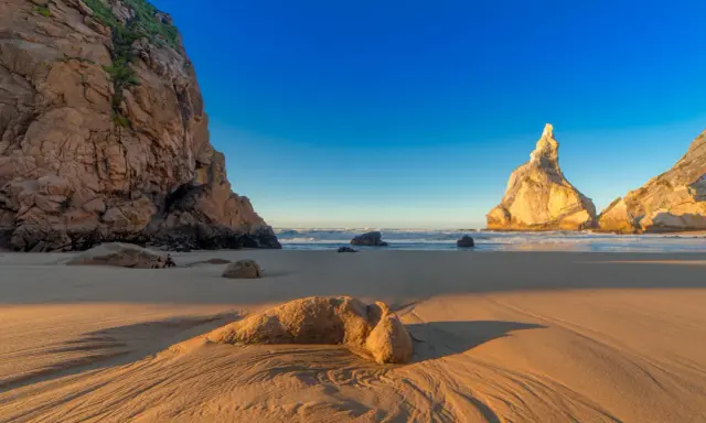 Goldene Sanddünen und Felsen am Praia da Ursa im warmen Abendlicht.