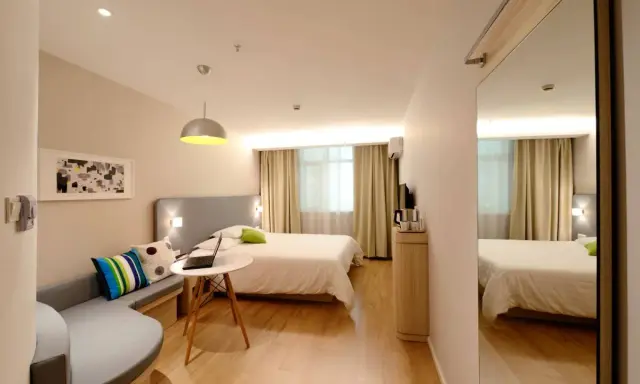 Habitación estrecha y alargada con cama, sofá curvo, mesa con portátil y espejo de cuerpo entero.