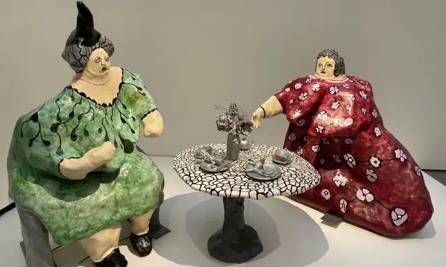 Dos figuras voluminosas, al estilo de Niki de Saint Phalle, sentadas a una mesa de té. Una viste de verde, la otra de rojo.