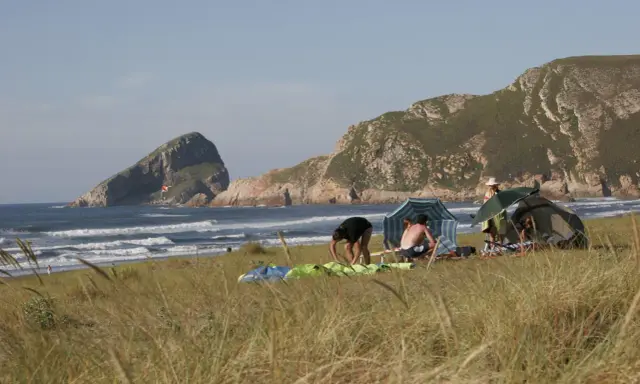 Gente en un playón de bayas, con carpas y sombrillas, disfrutando del mar y las montañas.