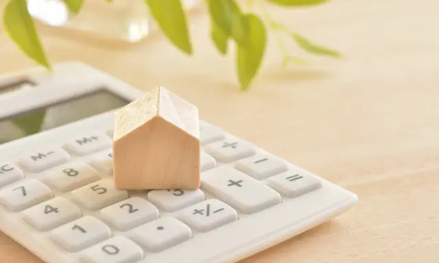 Maison en bois sur une calculatrice blanche, symbolisant le calcul du prix du **pel plafond**. Des feuilles vertes floues en arrière-plan.