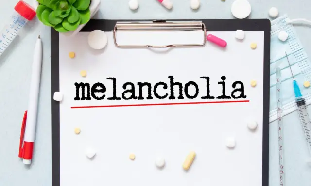 Na kartce widnieje napis "melancholia". Wokół leżą tabletki, strzykawka i maseczka, sugerując walkę z tym stanem.