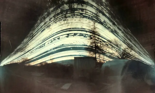 Paesaggio distorto con alberi e strutture, catturato con un **foro stenopeico**. Le strisce luminose suggeriscono un lungo tempo di esposizione.