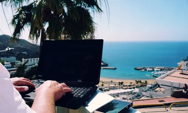 Praca na Cyprze: człowiek pisze na laptopie z widokiem na morze i palmę. Idealne miejsce na zdalną pracę.