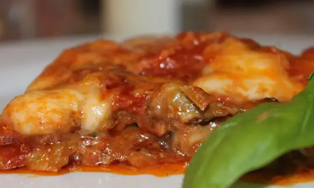 Deliciosa porción de parmigiana di melanzane, con capas de berenjena, salsa de tomate y queso fundido, adornada con una hoja de albahaca fresca.