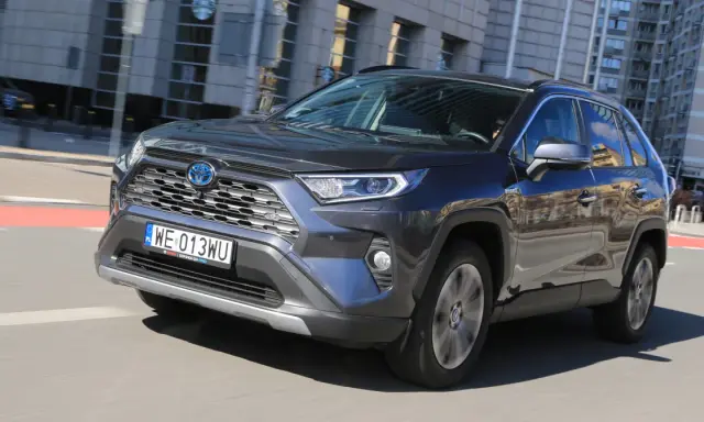 Ciemnoszary Toyota RAV4 Hybrid pędzi po mieście, odbijając w lustrach budynki i niebo.