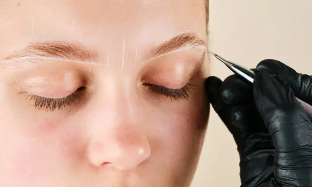 Une main gantée dessine des lignes blanches sur un sourcil pour un microblading poil à poil.