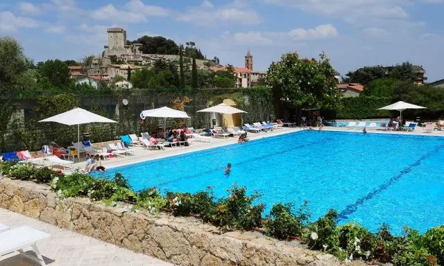 Piscina azzurra in un villaggio famiglie toscana, con castello medievale sullo sfondo e persone che si rilassano al sole.