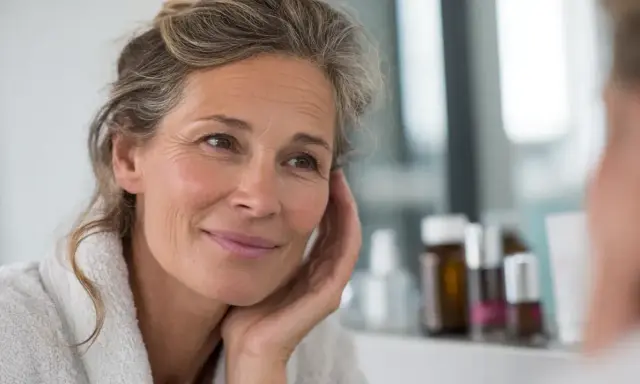 Une femme souriante, aux cheveux grisonnants, se regarde dans le miroir. Elle porte une robe de chambre douce, comme un **trèfle rouge** qui s'épanouit.