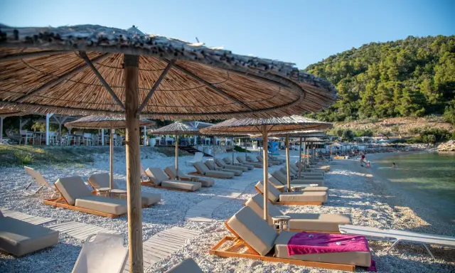 Alonissos spiagge: lettini e ombrelloni di paglia su una spiaggia di ciottoli, con il mare cristallino e la collina verde sullo sfondo.