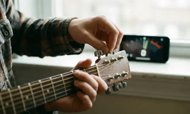 Main d'un musicien ajustant une guitare accordée, avec un accordeur sur un téléphone en arrière-plan.